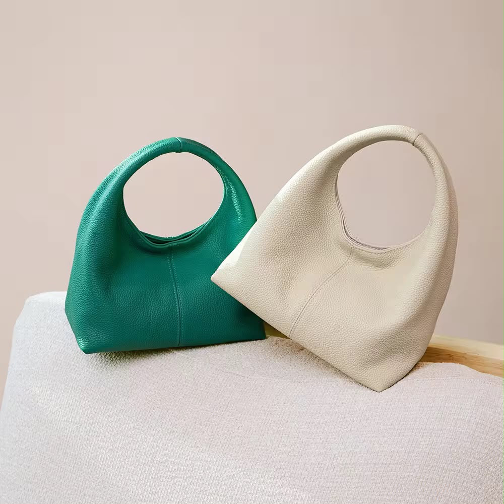 Two pebbled leather shoulder bags with circular handles, one in teal green and one in cream, displayed on a textured light beige surface against a solid beige background.