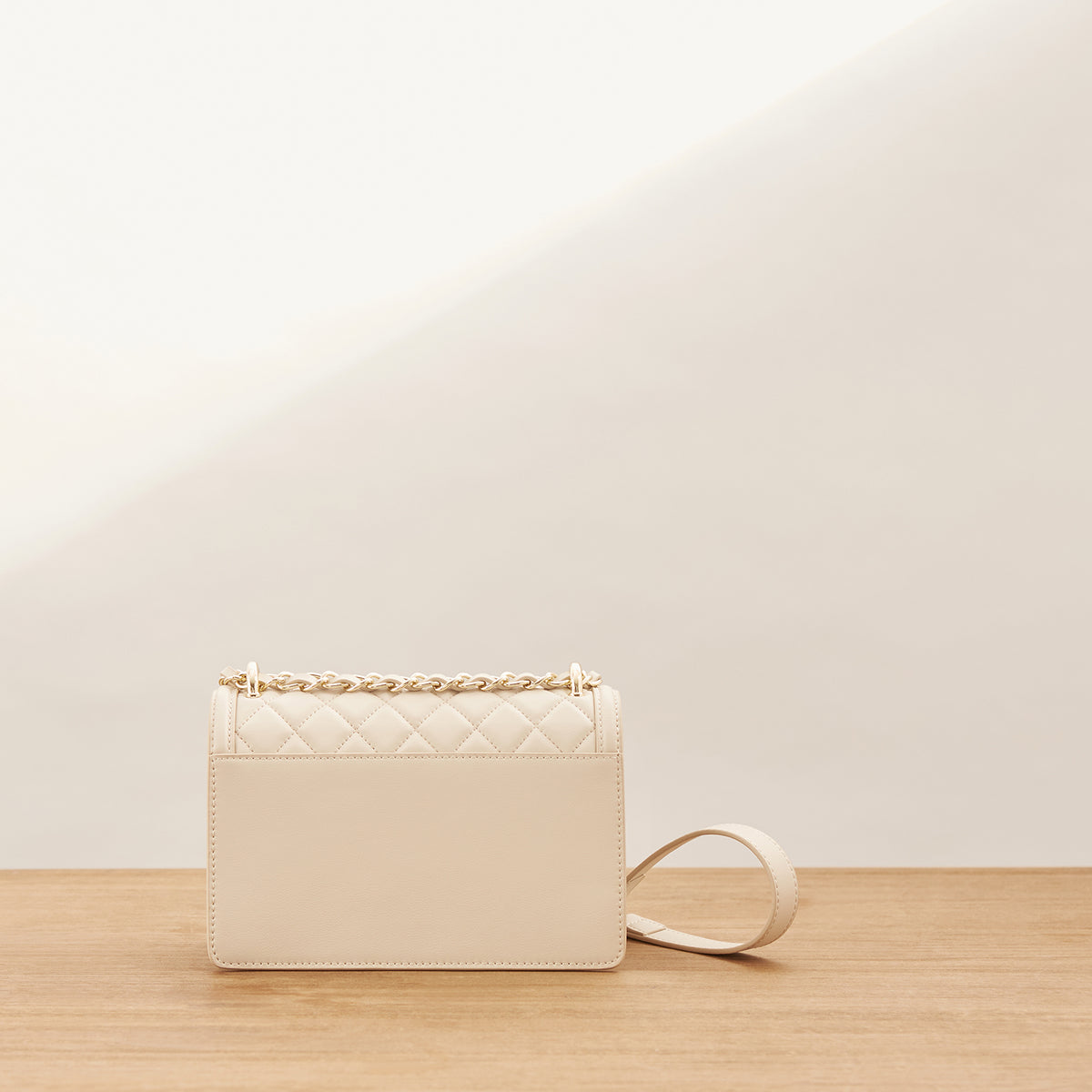 A cream-colored quilted leather shoulder bag with a gold chain handle, resting on a light wooden surface against a minimalist beige background.