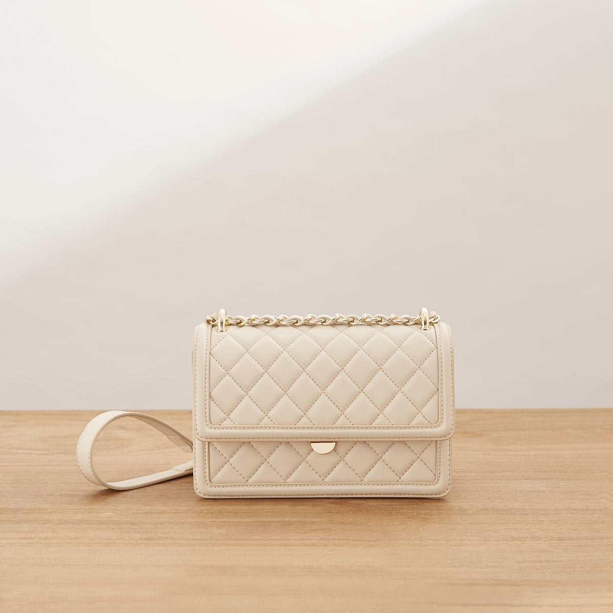 A cream-colored quilted leather shoulder bag with a gold chain strap and a small gold turn-lock clasp, resting on a light wood surface against a neutral beige background.